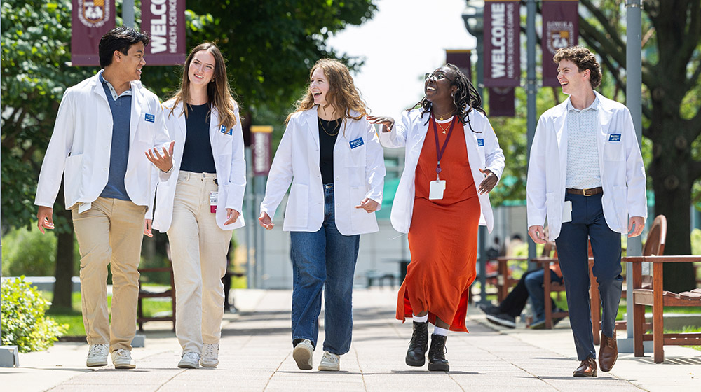 Five students in white coats walking and talking on campus